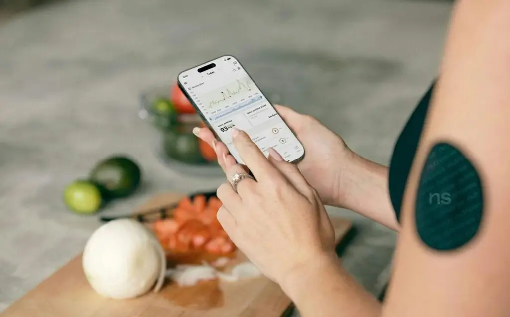 A woman using a smartphone app to track blood sugar levels with fresh vegetables in the background.